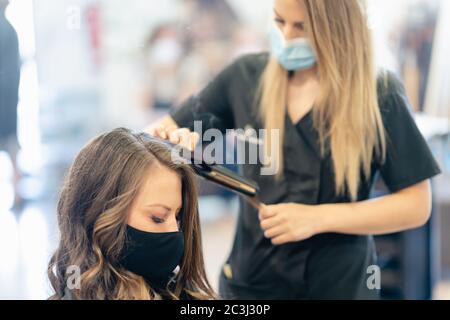 Friseurin, geschützt durch eine Maske, Kämmen der Haare ihres Kunden mit einem Haareisen in einem Salon. Stockfoto