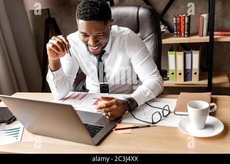 Lächelnder junger afrikanischer Geschäftsmann, der mit einem Kunden bei einem Videoanruf spricht, an einem Tisch im Büro sitzt Stockfoto