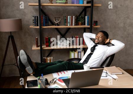 Lächelnder junger afrikanischer Geschäftsmann entspannt sich, während er an einem Tisch im Büro sitzt Stockfoto