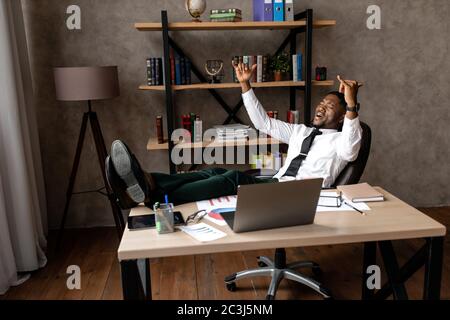 Lächelnder junger afrikanischer Geschäftsmann entspannt sich, während er an einem Tisch im Büro sitzt Stockfoto