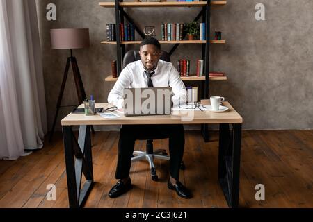 Erfolgreicher junger afrikanischer Geschäftsmann sitzt an seinem Schreibtisch im Büro und arbeitet auf einem Laptop Stockfoto