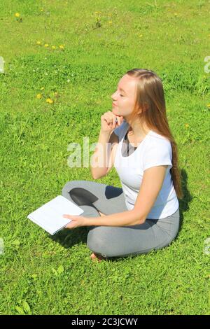 Ein junges, schönes Mädchen im weißen T-Shirt mit geschlossenen Augen sitzt auf dem grünen Gras auf dem Rasen mit einem Notizbuch und einem Stift, mit dem Handkinn gestützt, denkt und träumt, komponiert Text und Gedichte. Stockfoto