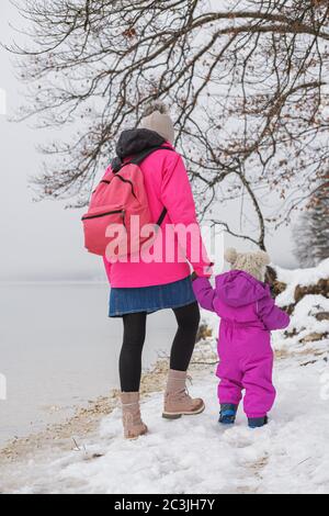 Blick von hinten auf eine Mutter und ihre Tochter, die auf Schnee am Wintersee spazieren gehen. Stockfoto