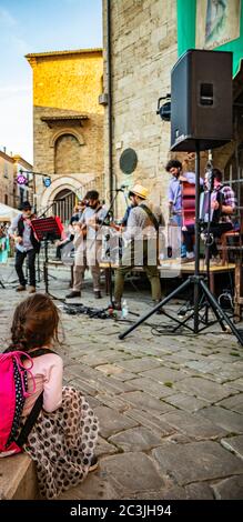 Bevagna, Perugia, Umbrien, Italien. Ein kleines Mädchen mit einem Zopf, bewundernswert, das eine Bandshow ansieht. Konzert von Straßenkünstlern während eines Dorffest. Stockfoto