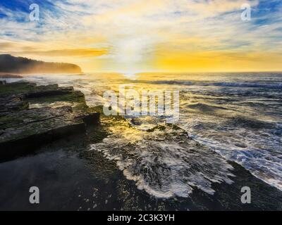 Wellen überfluten die Sandsteinplatte am Turimetta Strand der Pazifikküste von Sydney im hellen aufgehenden Sonnenlicht. Stockfoto