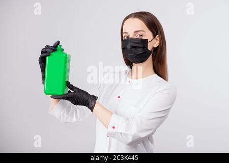 Desinfektionsmittel in grünem Glas, Mädchen in Maske und Handschuhe hält ein Antiseptikum in den Händen, hellgrauer Hintergrund Stockfoto