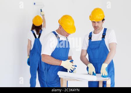 Arbeiter in blauer Uniform und gelben Harthüten mit Blaupause arbeiten auf der Baustelle Stockfoto