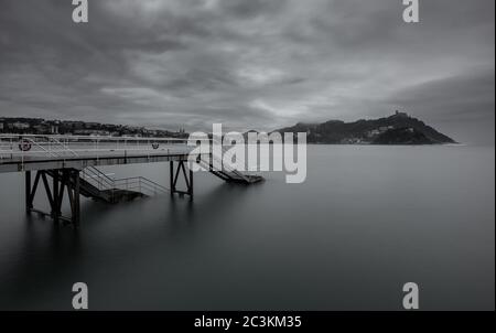 Graustufenaufnahme einer hölzernen Promenade am Meer unter dunklen Sturmwolken in San Sebastian, Spanien Stockfoto