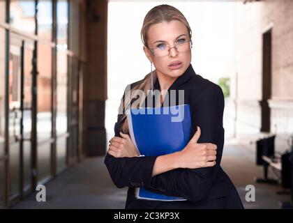 Geschäftsfrau in Brillen in einem Business-Anzug hält fest einen Ordner mit wichtigen Dokumenten und ungläubig schaut auf die Kamera. Stockfoto