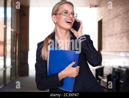 Eine Frau in einem Business-Anzug trägt einen Ordner mit Dokumenten und lächelt, während sie am Telefon spricht. Geschäftskonzept. Stockfoto