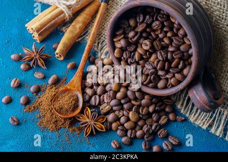 Kaffeesorten, die aus einer Tonschale ausströmen und auf einem blauen texturellen Hintergrund verstreut sind, Anissterne, Zimtstangen und gemahlener Kaffee in einem Holzlöffel Stockfoto
