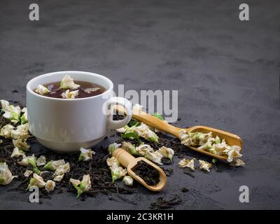 Schwarzer Tee in einer weißen Tasse auf einem texturellen Hintergrund, mit trockenem Tee bestreut um und trockenen Jasminblüten Stockfoto