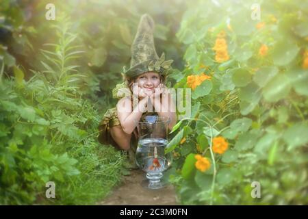 Lustiges Mädchen in einem Gnom Hut und einem Anzug in einem grünen Garten, sitzt neben einem Aquarium mit einem Goldfisch. Märchen. Stockfoto