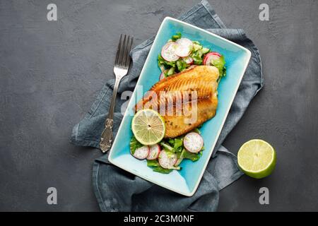 Gegrillter Fisch Seebarsch mit frischen Kräutern, Rettich und Limette in einem rechteckigen blauen Teller. Dunkler Hintergrund. Stockfoto