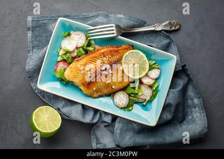 Gegrillter Fisch Seebarsch mit frischen Kräutern, Rettich und Limette in einem rechteckigen blauen Teller. Dunkler Hintergrund. Stockfoto