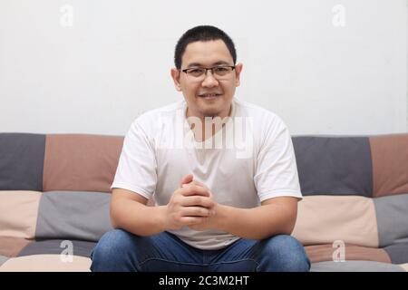 Schöner junger asiatischer Mann, der zu Hause auf dem Sofa sitzt, glücklich lächelt und fröhlich auf die Kamera schaut Stockfoto