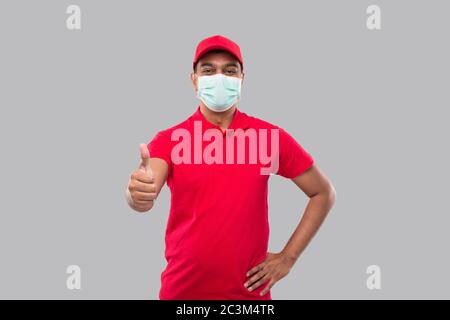 Delivery Man Zeigt Daumen Nach Oben Tragen Von Medizinischen Maske Isoliert. Indische Lieferung Junge Lächelnd. Lieferung Nach Hause. Stockfoto