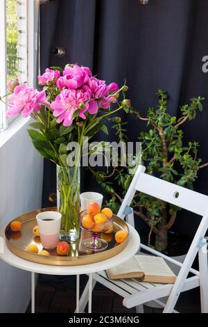 Sommer-Frühstück auf dem Balkon. Zwei Tassen Getränk, Bouquet von rosa Pfingstrosen, geöffnetes altes Buch und frisches Obst auf weißem Tisch. Stockfoto