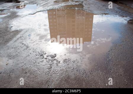 Die schlechte asphaltierte Straße mit einem großen Schlagloch gefüllt mit Wasser. Gefährlich zerstörte Straßenbett. Spiegelreflexion eines Gebäudes im Wasser. Stockfoto