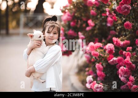 Lächelndes Baby Mädchen hält Haustier Hund über Natur Hintergrund aus nächster Nähe. Blick auf die Kamera. Selektiver Fokus auf Welpen. Stockfoto