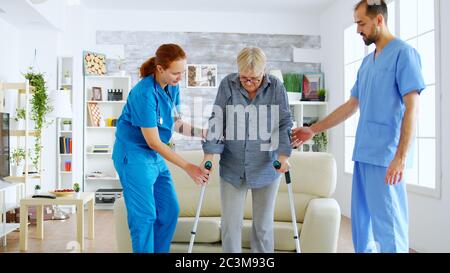 Ältere Frau mit Krücken bekommen Hilfe für weibliche Krankenschwester und Arzt auf ihren Beinen von der Couch stehen Stockfoto