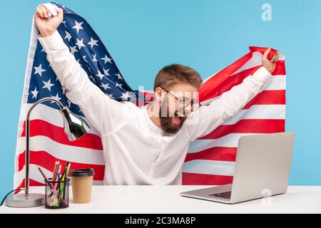 Glücklicher Geschäftsmann, der die amerikanische Flagge hochhebt, live patriotisches Programm auf dem Laptop am Arbeitsplatz beobachtet und vor Freude schreit, den 4. Tag der US-Unabhängigkeit zu feiern Stockfoto