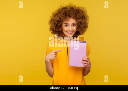 Portrait einer positiven lockigen Frau im urbanen Stil Hoodie, der auf ein leeres Notizbuch zeigt, Werbefläche auf Papier, Platz für To-Do-Liste, Idee. Stockfoto
