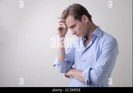 Junger Mann mit einem blauen Hemd, der tiefe Gedanken im Stehen hat Gegen eine weiße Wand Stockfoto