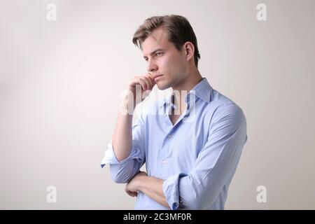 Junger Mann mit einem blauen Hemd, der tiefe Gedanken im Stehen hat Gegen eine weiße Wand Stockfoto