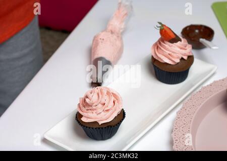 Schokoladencupcakes mit Erdbeercreme. Dekoriert mit halben Erdbeeren in Schokolade. Neben den Rohlingen und Zutaten für Cupcakes. Stockfoto