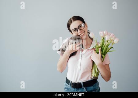 Studio Portrait von jungen Erwachsenen hübsches Mädchen mit Brille in Rosa T-Shirt gekleidet hält Bouquet von frischen Tulpen und ihre kleine Katze Stockfoto