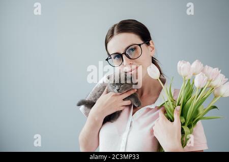 Studio Portrait von jungen Erwachsenen hübsches Mädchen mit Brille in Rosa T-Shirt gekleidet hält Bouquet von frischen Tulpen und ihre kleine Katze Stockfoto