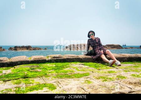 Eine glückliche chinesin, die an einem sonnigen Tag in der Nähe der felsigen Küste der Fushan Bay in der chinesischen Provinz qingdao shandong sitzt. Stockfoto