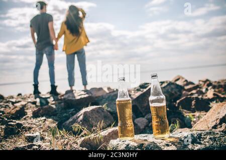 Nahaufnahme zwei Flaschen Bier stehen auf Steinen in der Nähe des Wassers in der Sonne auf einem Hintergrund eines Paares. Mann und Frau halten die Hände. Stockfoto