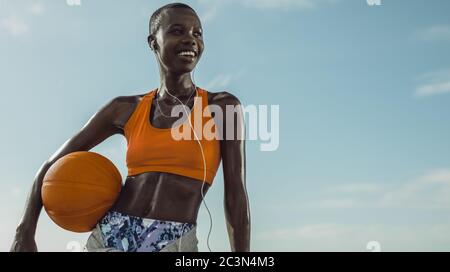 Frau in Fitness tragen im Freien stehend hält einen Basketball. Basketballspielerin schaut weg und lächelt gegen den Himmel.