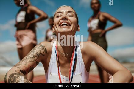 Läufer sitzt auf der Strecke mit Medaille nach dem Sieg und lächeln. Der Sieger des Laufrennens sitzt auf der Strecke mit Athleten im Hintergrund. Stockfoto