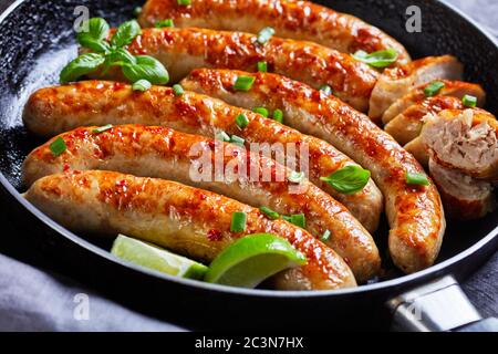 Nahaufnahme einer gebratenen Schweinswurst-Glieder aus Hackfleisch auf einer Pfanne mit frischem Basilikum und Frühlingszwiebeln auf der Oberseite, Kalk Keile, auf einem dunklen Holztisch serviert Stockfoto