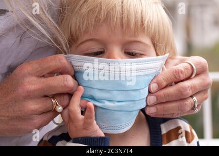 Mutter gibt einem überraschten kleinen Kind Kleinkind medizinische Gesichtsmaske auf zum Schutz gegen Coronavirus Covid-19 Pandemie. Stockfoto