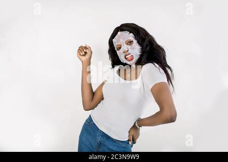 Gesichtspflege und Schönheitsbehandlungen. Lächelnd hübsche afrikanische Frau mit einem Tuch Papier Blatt feuchtigkeitsspendende Maske auf ihrem Gesicht, posieren und Spaß haben, isoliert Stockfoto