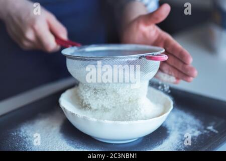 Ein Mann siebt krümeliges Weizenmehl durch ein Metallsieb in eine weiße Schüssel. Garvorgang. Stockfoto