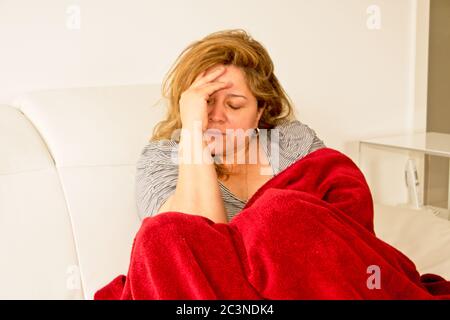 Blonde Frau, die auf einem Sofa liegt, mit der Hand auf dem Kopf, mit einem depressiven Ausdruck und einer roten Decke über ihr Stockfoto