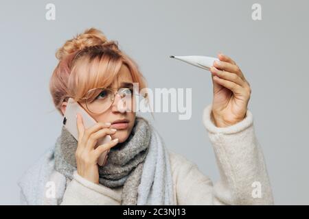 Nahaufnahme Porträt der europäischen ungesunden Frau in Brille kalt erwischt und Blick auf Thermometer. Krank junge blonde Frau im Schal Gefühl schlecht, reden Stockfoto