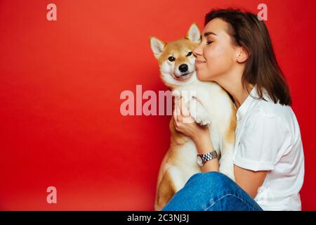 Nette Brünette Frau in weißen T-Shirt und Jeans halten und umarmen Shiba Inu Hund auf Ebene roten Hintergrund. Liebe zu den Tieren, Haustiere Konzept Stockfoto