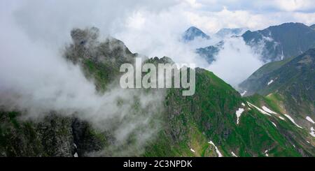 Hohe Berggipfel zwischen den Wolken. Wunderschöne Sommerlandschaft von fagaras Grat mit etwas Schnee auf Hügeln. wolkiges Wetter mit blauem Himmel Stockfoto