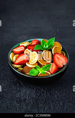 Trendiges Frühstück mit winzigen Pfannkuchen (Mini-Pfannkuchen) mit Erdbeeren und Minze in einer Schüssel (Tasse) auf dunklem Hintergrund. Hintergrund Für Das Essen. Mini-Cerealien Stockfoto