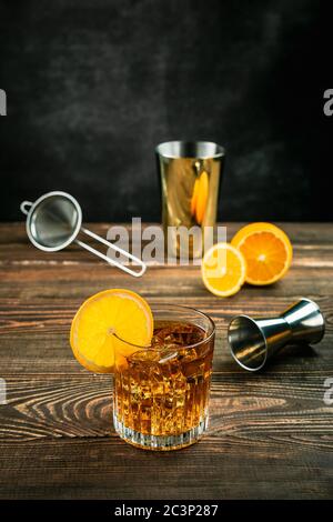 Ein oranger alkoholischer Cocktail in einem mit Eis gefüllten Steinglas, garniert mit einem orangen Rad. Eine geschnittene Orange, Jigger, Shaker und Sieb. Holzta Stockfoto