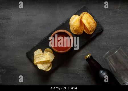 Glas und Flasche Bier mit Chips und Tomatenketchup auf dunklem Steingrund Stockfoto