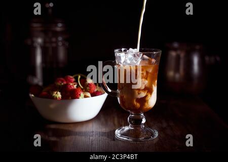 Eiskaffee in einem hohen Glas mit übergossenem Sahne, Eiswürfeln und Bohnen auf einem alten rustikalen Holztisch mit einem Erdbeerplatte im Hintergrund. Stockfoto