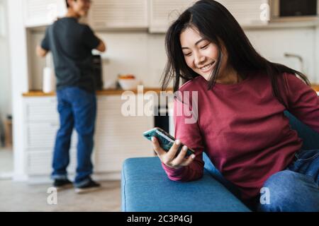 Lächelnde junge asiatische Frau mit Handy, während sie zu Hause auf einer Couch mit ihrem Mann auf einem Hintergrund sitzt Stockfoto