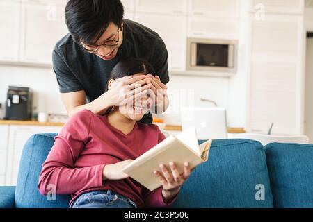 Foto von freudigen asiatischen Mann bedeckend die Augen der Freundin, während sie Buch auf dem Sofa in gemütlichen Zimmer zu lesen Stockfoto